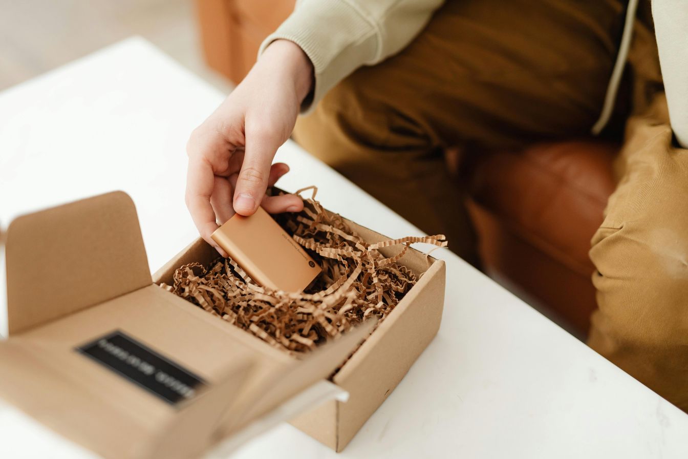 Person unpacking a small electronic device from a cardboard box with shredded paper filler, showing eco-friendly protective packaging.