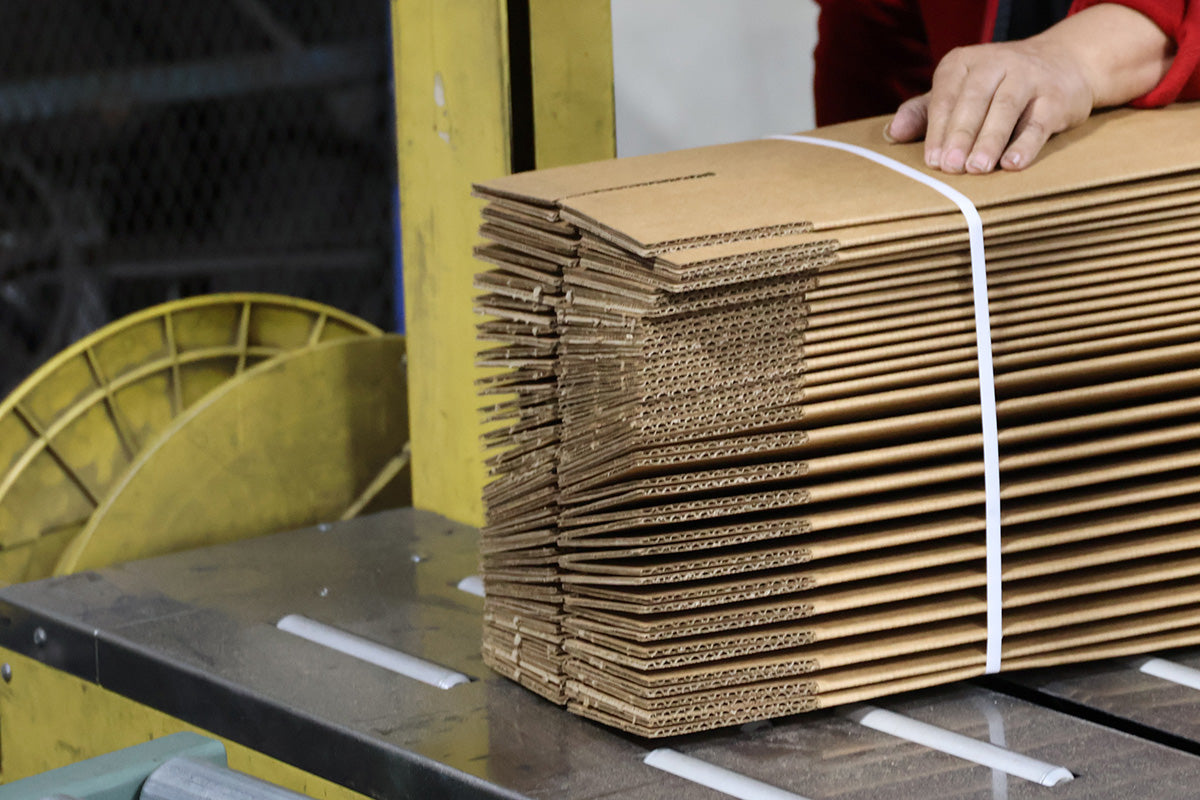 Stack of flat cardboard boxes tied with a white strap on a production line, ready for packaging or delivery.