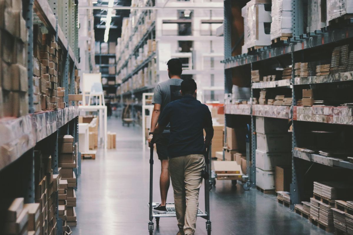 Two people in a warehouse pushing a cart through aisles filled with stacked cardboard boxes, representing logistics and packaging storage.