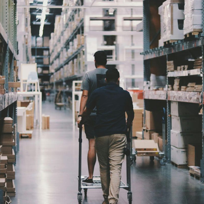 Two people in a warehouse pushing a cart through aisles filled with stacked cardboard boxes, representing logistics and packaging storage.