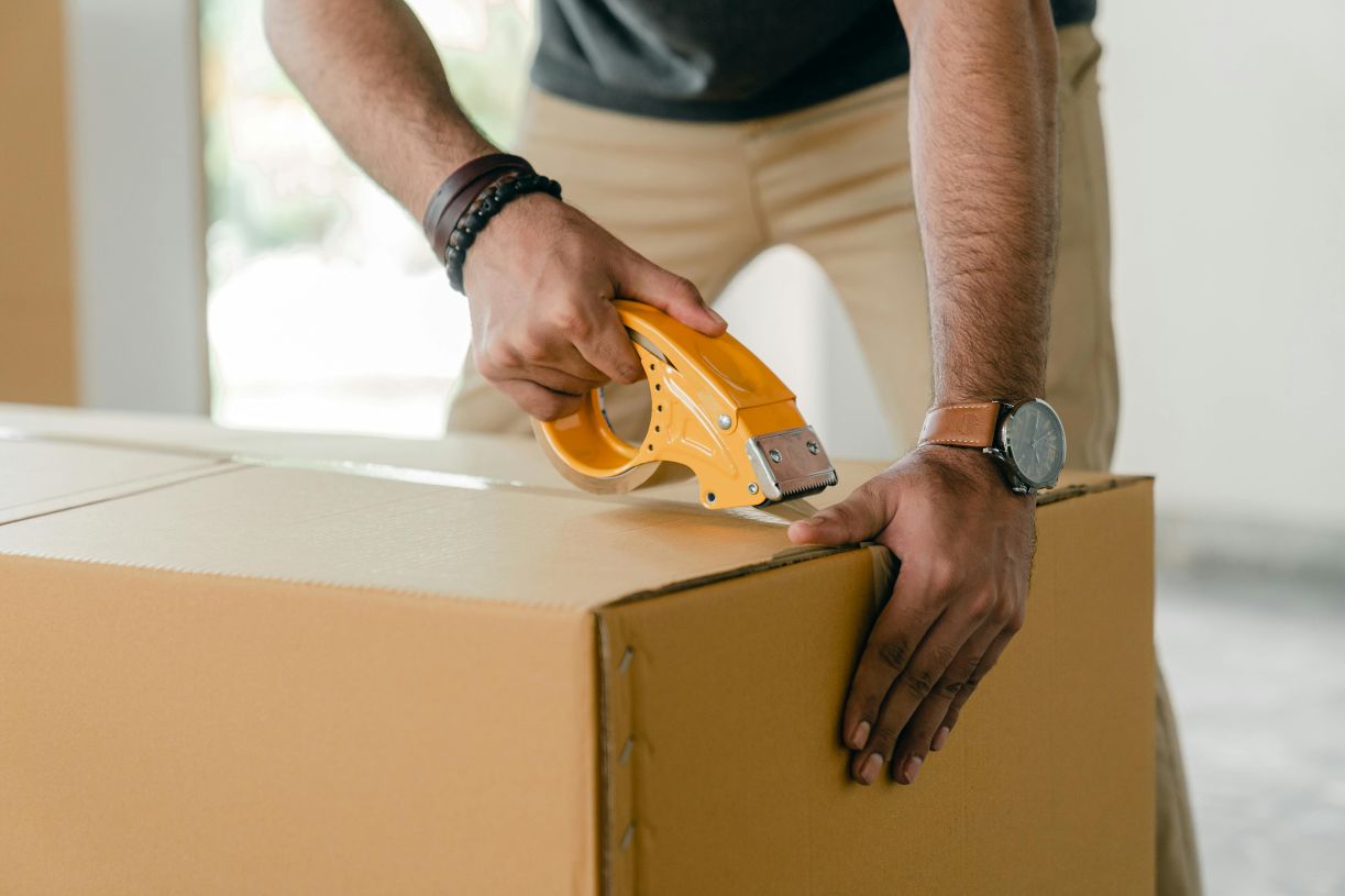 Person sealing a cardboard box with packing tape using a tape dispenser, representing packaging and shipping preparation.
