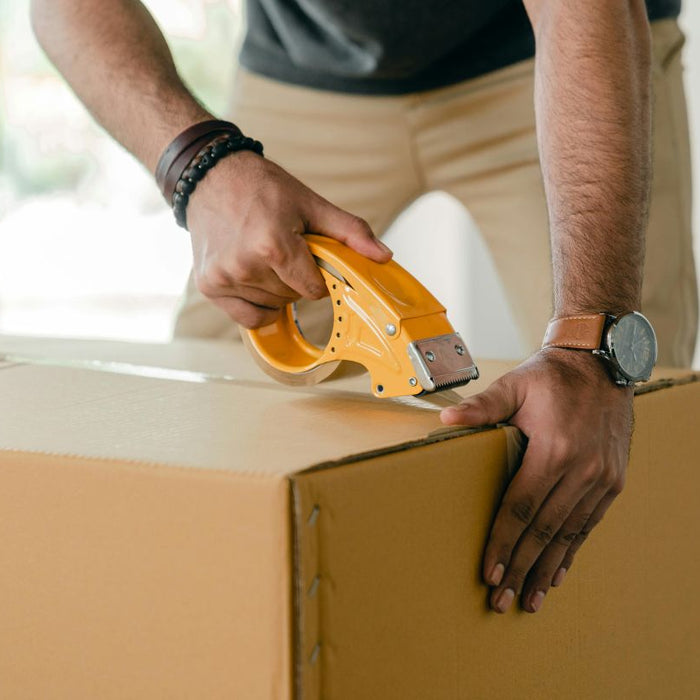 Person sealing a cardboard box with packing tape using a tape dispenser, representing packaging and shipping preparation.