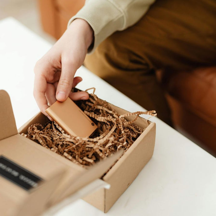 Person unpacking a small electronic device from a cardboard box with shredded paper filler, showing eco-friendly protective packaging.
